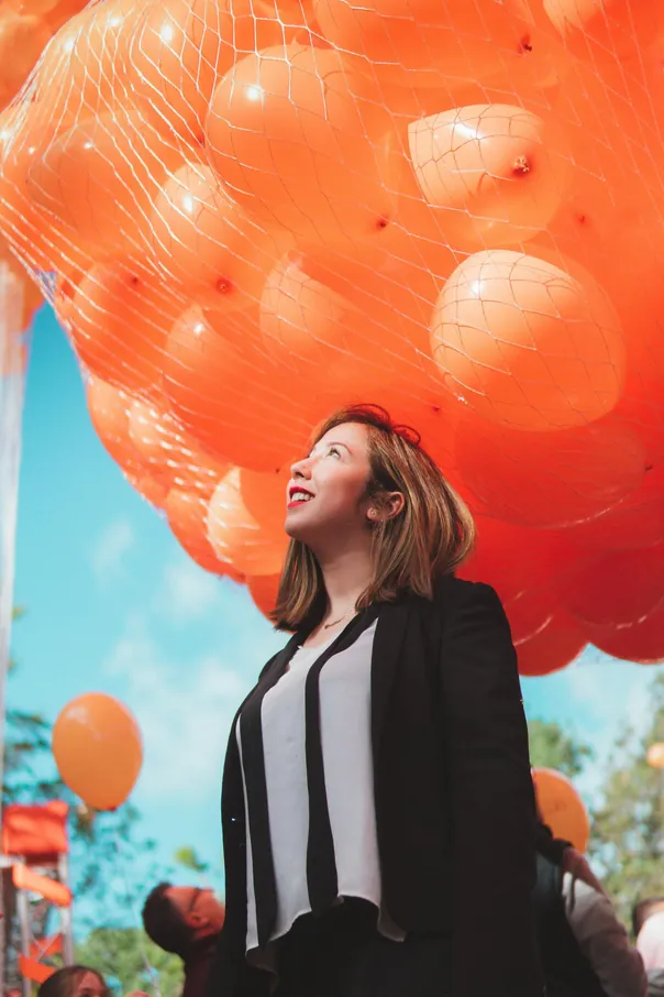 Young woman smiling under a display of vibrant orange balloons outdoors. Festive and joyful atmosphere.